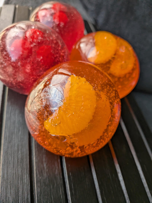 May include: Four spherical soaps with a translucent finish. Two soaps are red and contain embedded red berries, while the other two are orange and contain embedded orange slices. Each soap features visible air bubbles throughout the clear material.